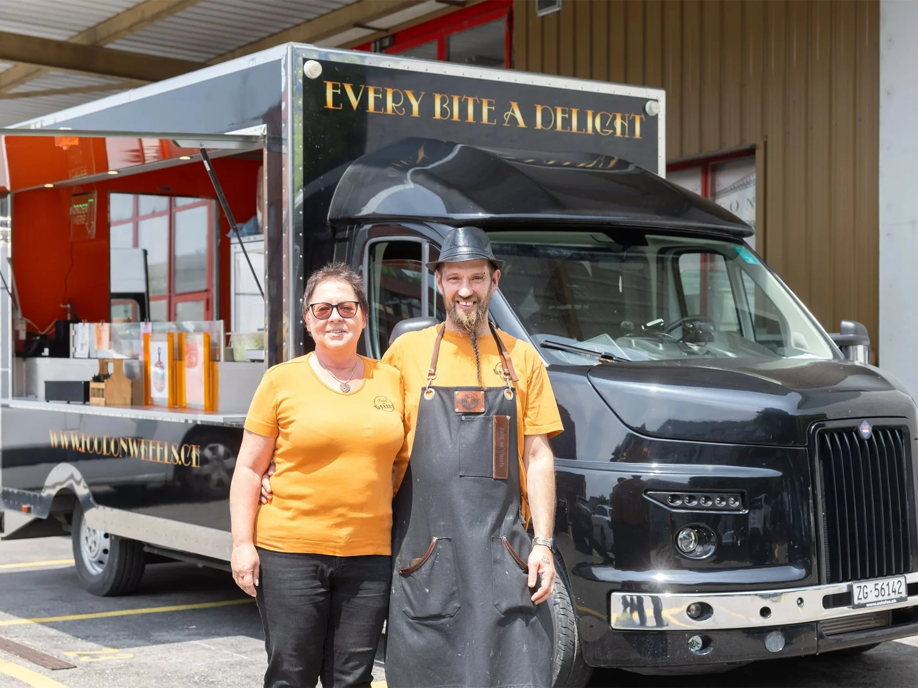 Claudia und Enrico Tezzele in orangen Food on Wheels Shirts vor ihrem schwarzen Foodtruck